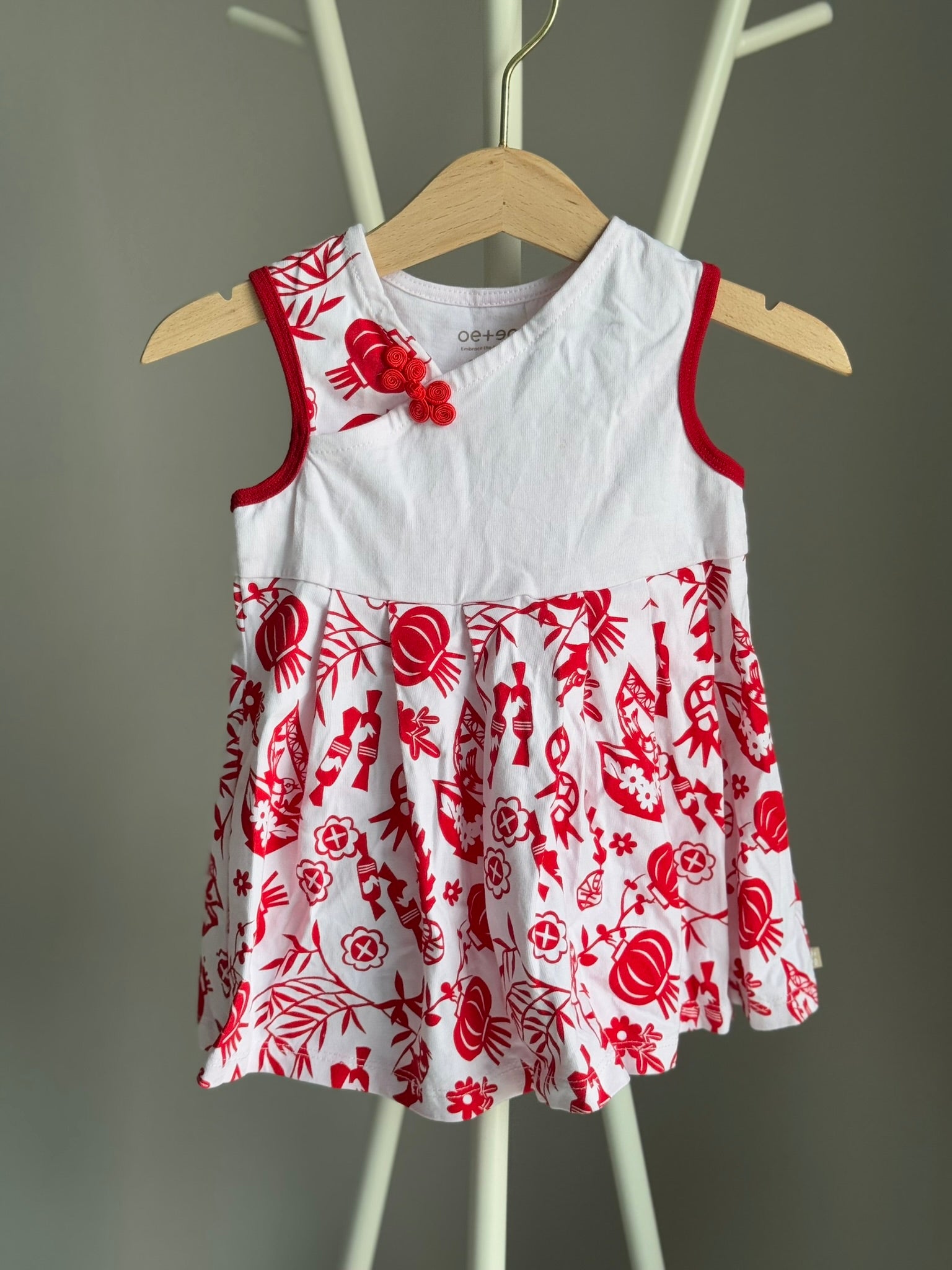 A red and white baby cheongsam dress with paper cutting patterns, displayed on a wooden hanger.