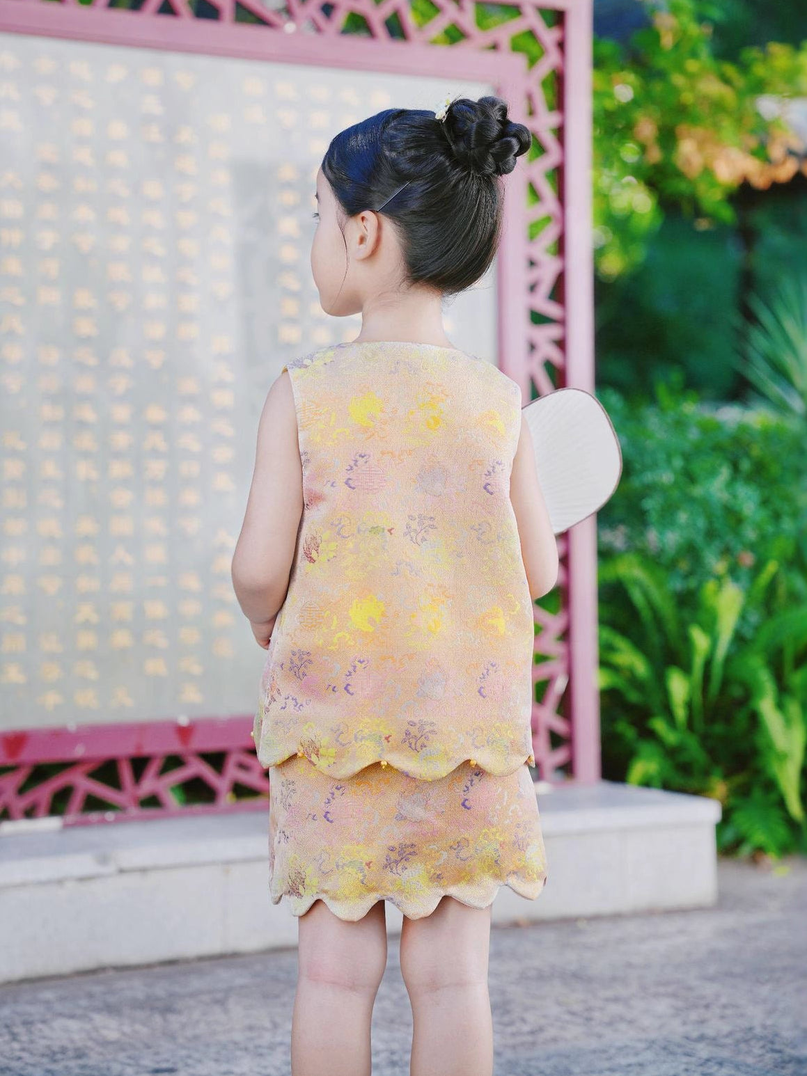 Child in a yellow dress standing in front of a decorative pink archway with greenery.