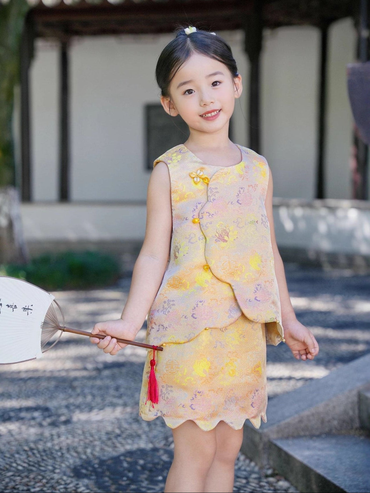 Young girl in a yellow floral dress holding a white parasol with a red tassel in an outdoor setting.