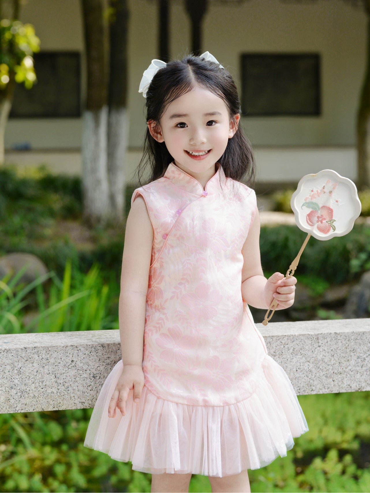 A young girl wearing a pink sleeveless cheongsam tulle dress with peach blossom design, holding a fan and standing by a tree.