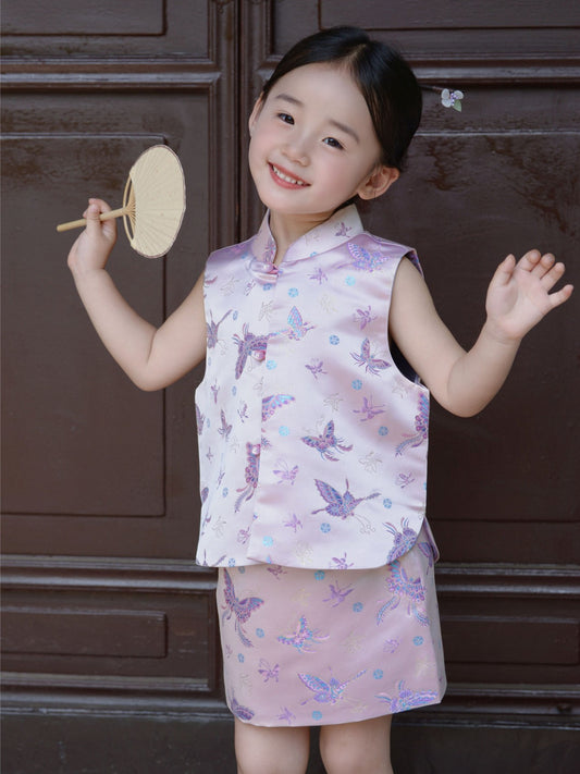 A young girl wearing a traditional purple cheongsam vest with a butterfly pattern and holding a fan, standing in front of a dark door.